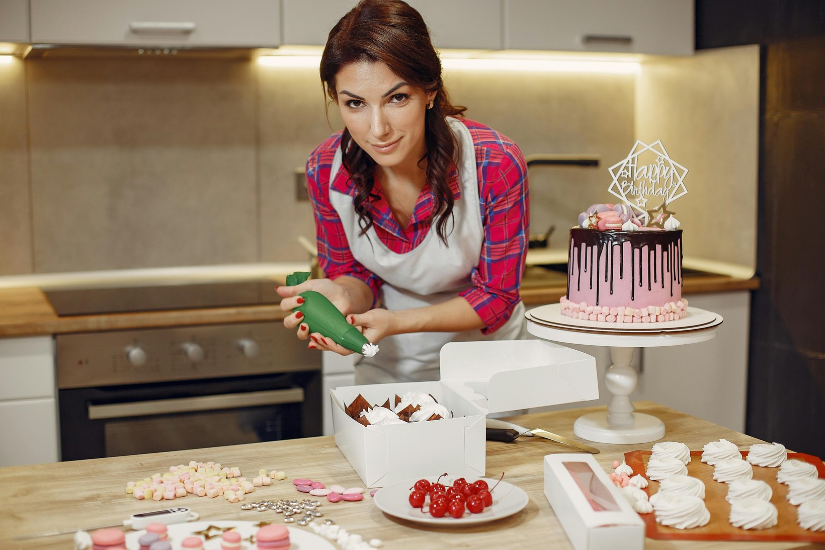 Woman in an apron piping icing onto chocolates in a kitchen with a decorated birthday cake on a stand beside her