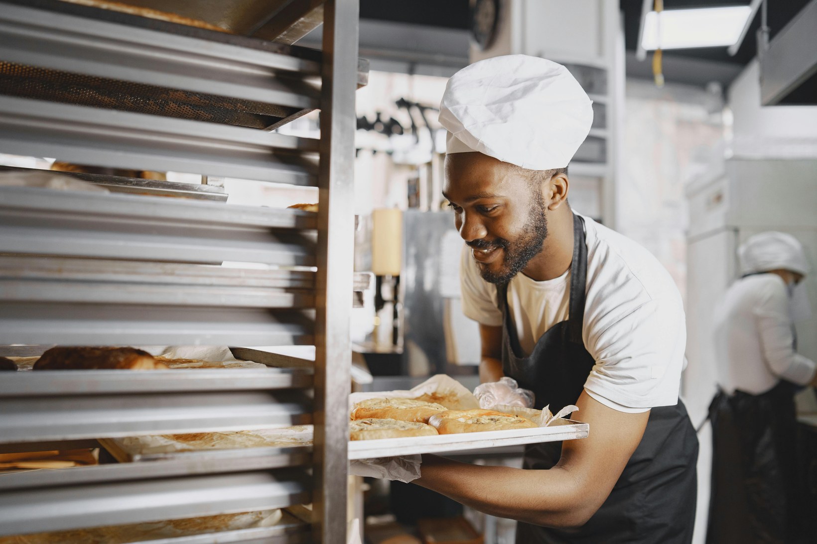 Baker in a chef's hat loading a tray of fresh rolls into a rack in a commercial bakery kitchen