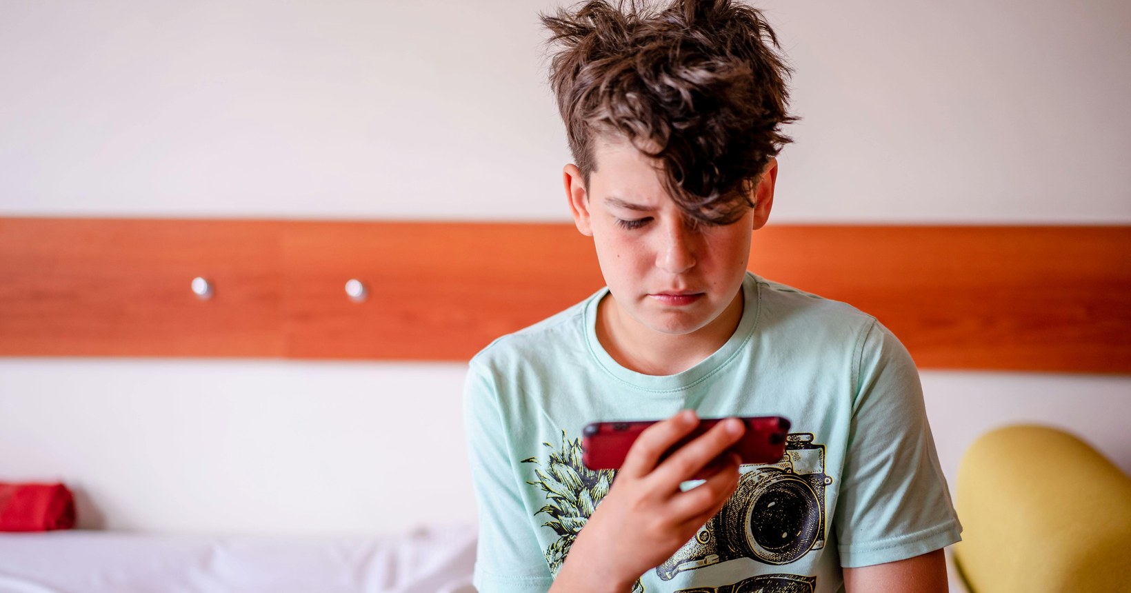 Young boy looking intently at a smartphone while sitting on a bed, representing younger generations' relationship with technology