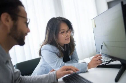 Woman pointing at a row of data on a computer monitor while a colleague looks on